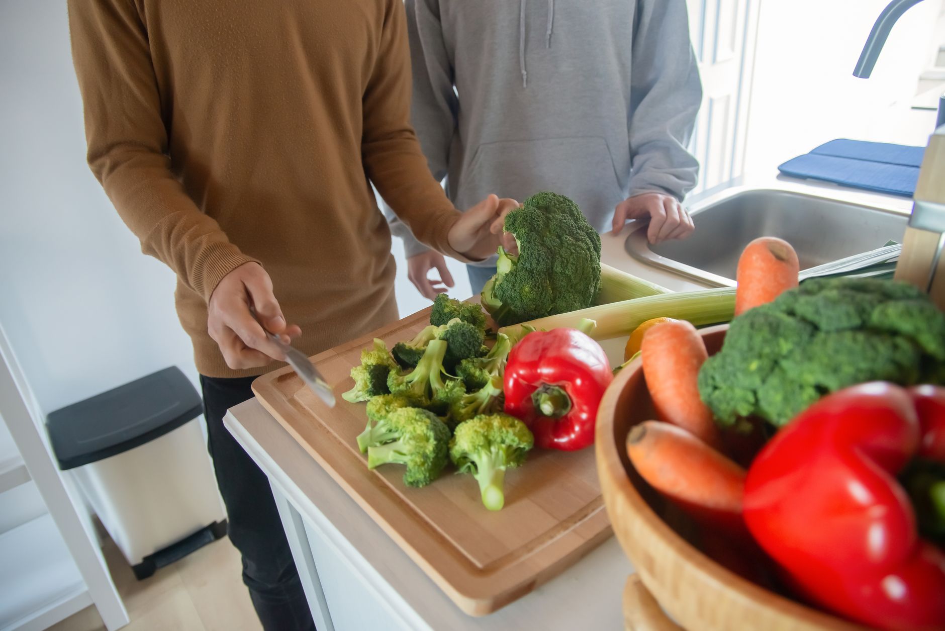 Deux personnes préparent des légumes dans une cuisine : brocoli, poivron rouge et carottes sur une planche et dans un bol en bois.