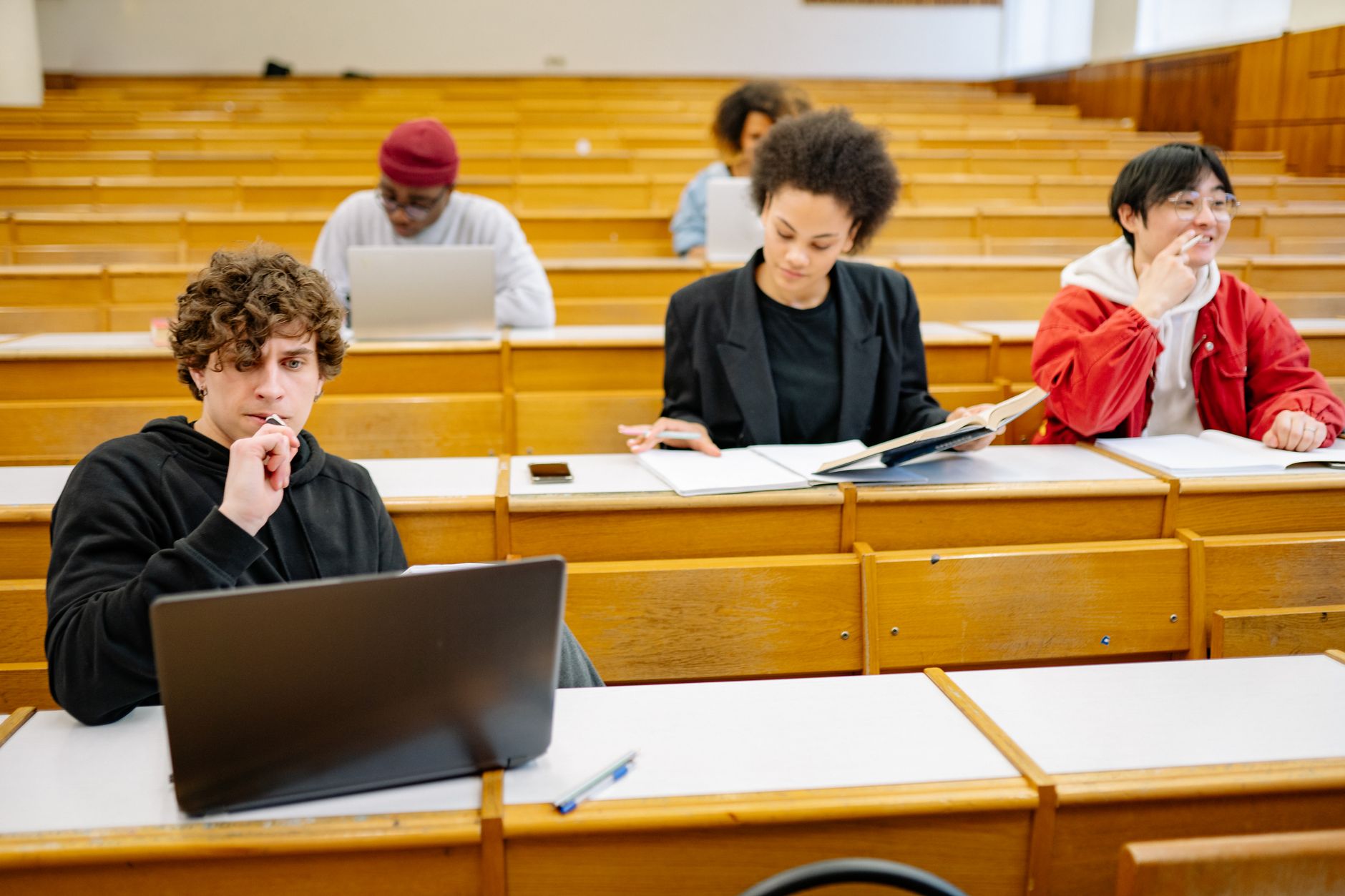 Étudiants dans un amphithéâtre aux bancs en bois, certains devant des ordinateurs portables, d’autres lisant ou prenant des notes.
