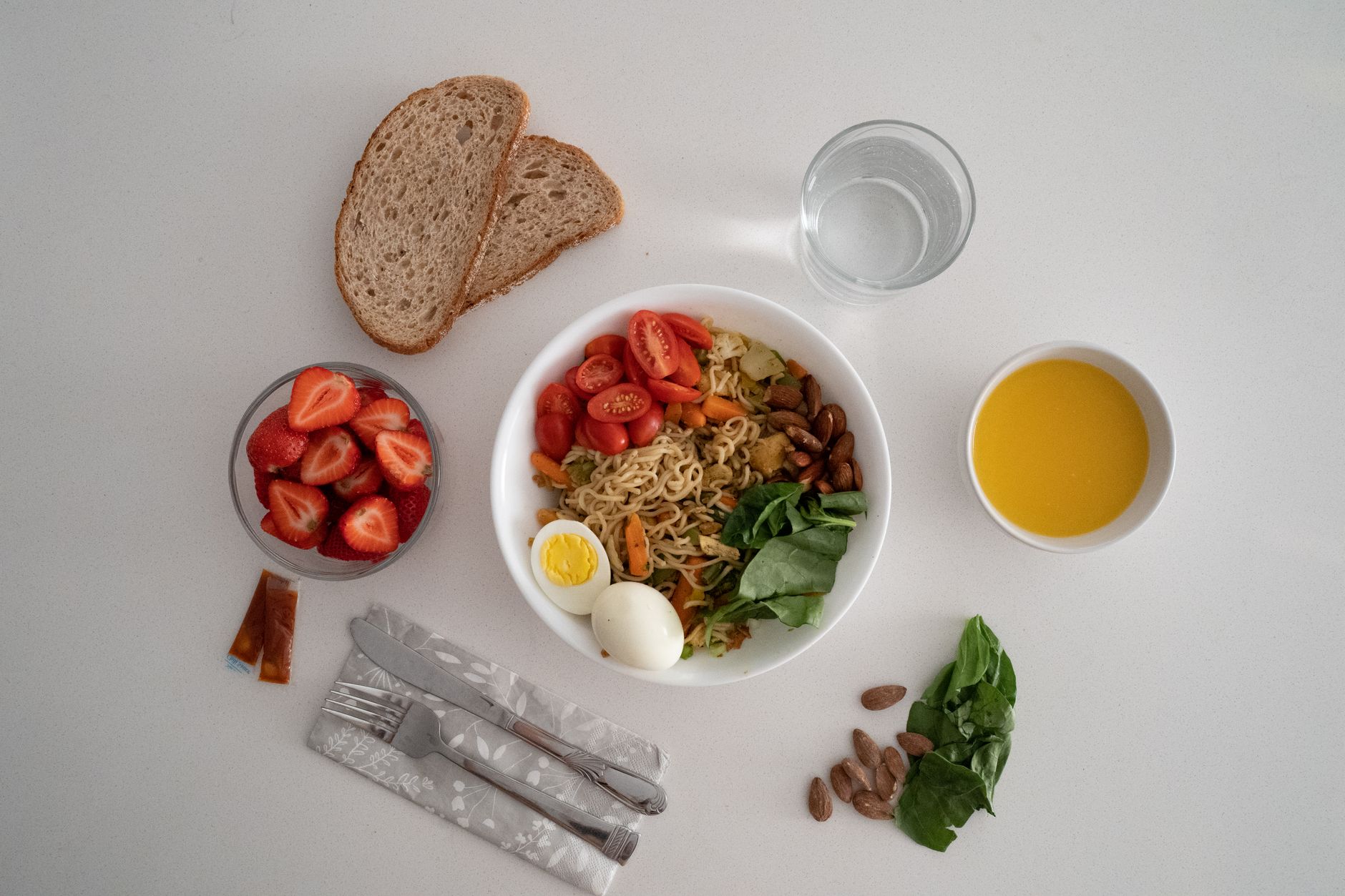Repas vu de dessus : bol de nouilles aux légumes et œuf dur, pain complet, fraises, verre d’eau, amandes et épinards sur une table blanche.