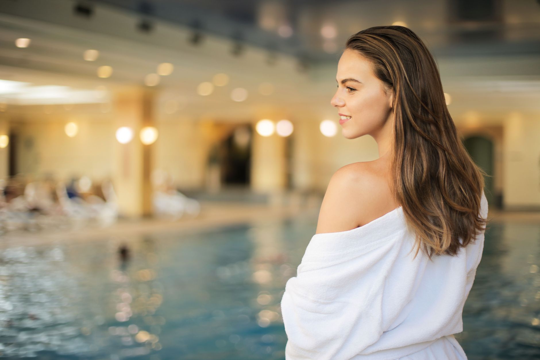 Une femme en peignoir blanc, de profil, sourit devant une piscine intérieure dans un cadre de spa aux lumières chaleureuses.